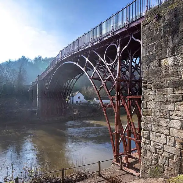 Uma imagem realista da Iron Bridge, a primeira ponte de ferro do mundo, construída em 1779 sobre o rio Severn, na Inglaterra. A cena mostra a ponte arqueada em ferro fundido, com detalhes das estruturas rebitadas e o arco principal refletido na água tranquila do rio. O fundo inclui colinas verdes e algumas construções de época ao redor, criando uma atmosfera histórica. Luz suave de fim de tarde, céu levemente nublado. Perspectiva de leve ângulo lateral para valorizar a arquitetura.